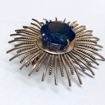 Vintage sunburst brooch with rose gold rays radiating from a round, deep blue alexandrite gemstone at the center, creating a striking purple and blue effect against the gold-toned metal.