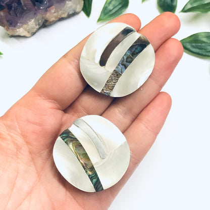 Vintage abalone shell clip-on earrings with abstract mother of pearl inlay design, held in palm of hand against white background with greenery.