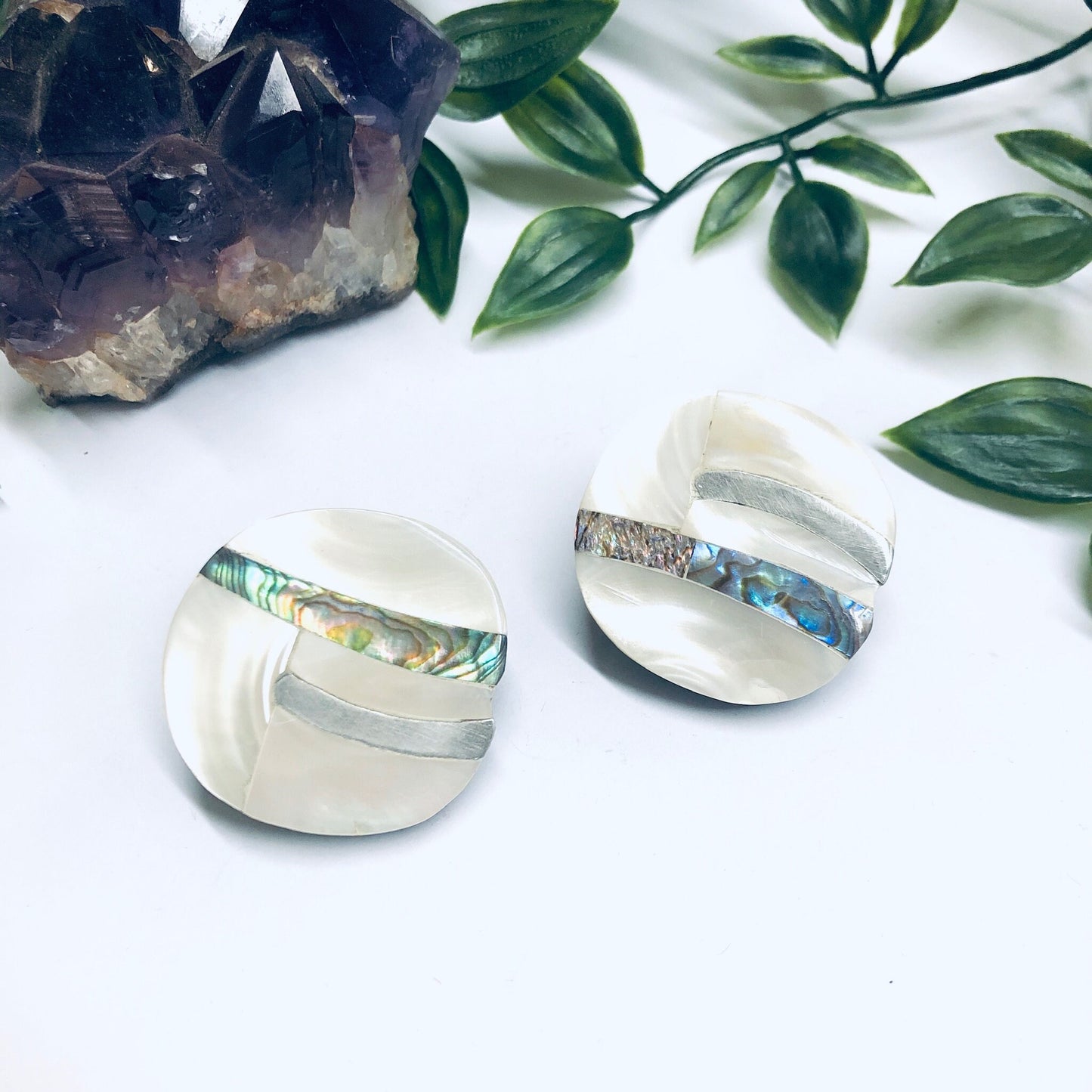 Vintage abalone shell clip-on earrings with abstract mother of pearl inlay design, displayed with dark crystal and green leaves on white marble background.