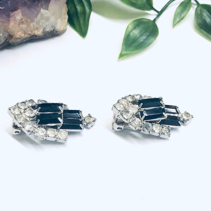 Sparkling black and silver rhinestone clip-on earrings next to crystal and green plant leaves on white background, retro fashion accessory