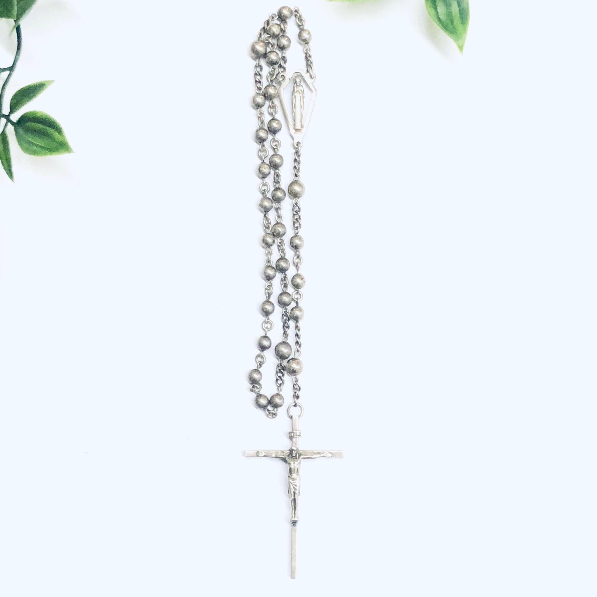 Silver rosary prayer beads with crucifix pendant on white background surrounded by green leaves, Catholic religious jewelry