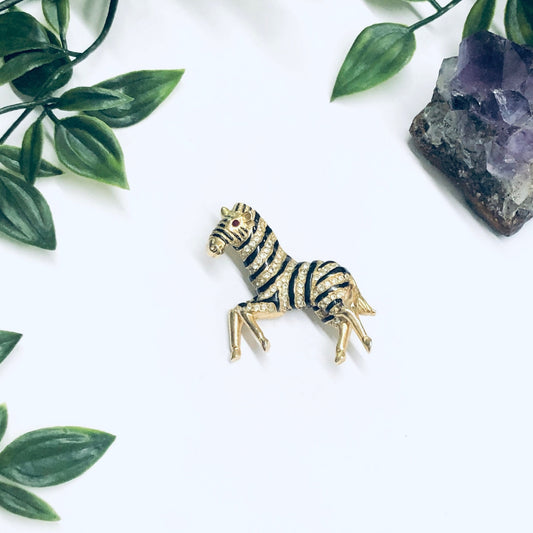 Gold-toned rhinestone zebra brooch pin surrounded by green foliage and amethyst crystal on white background