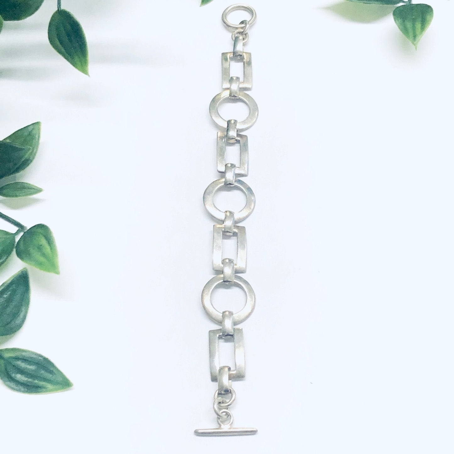 Geometric silver link bracelet with circle and rectangle cutouts and toggle closure, shown with green leaf accents