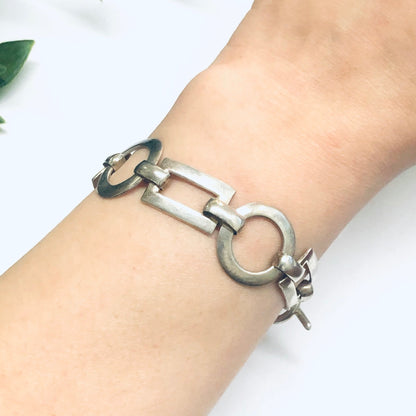 Silver geometric link bracelet with circle and rectangle cutouts, featuring a toggle closure clasp, shown on a person's wrist next to a green leaf.