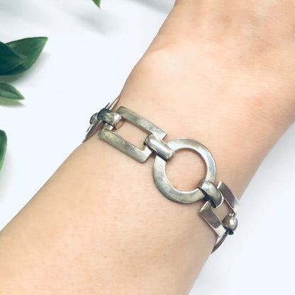 Silver geometric link bracelet with toggle closure, featuring alternating circle and rectangle shaped links, shown on a wrist against a white background with green leaves.