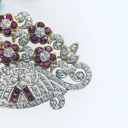 Vintage 18K yellow gold brooch with ruby and diamond floral design, featuring intricate scrollwork accented by round and marquise-cut diamonds and vibrant ruby flower buds.