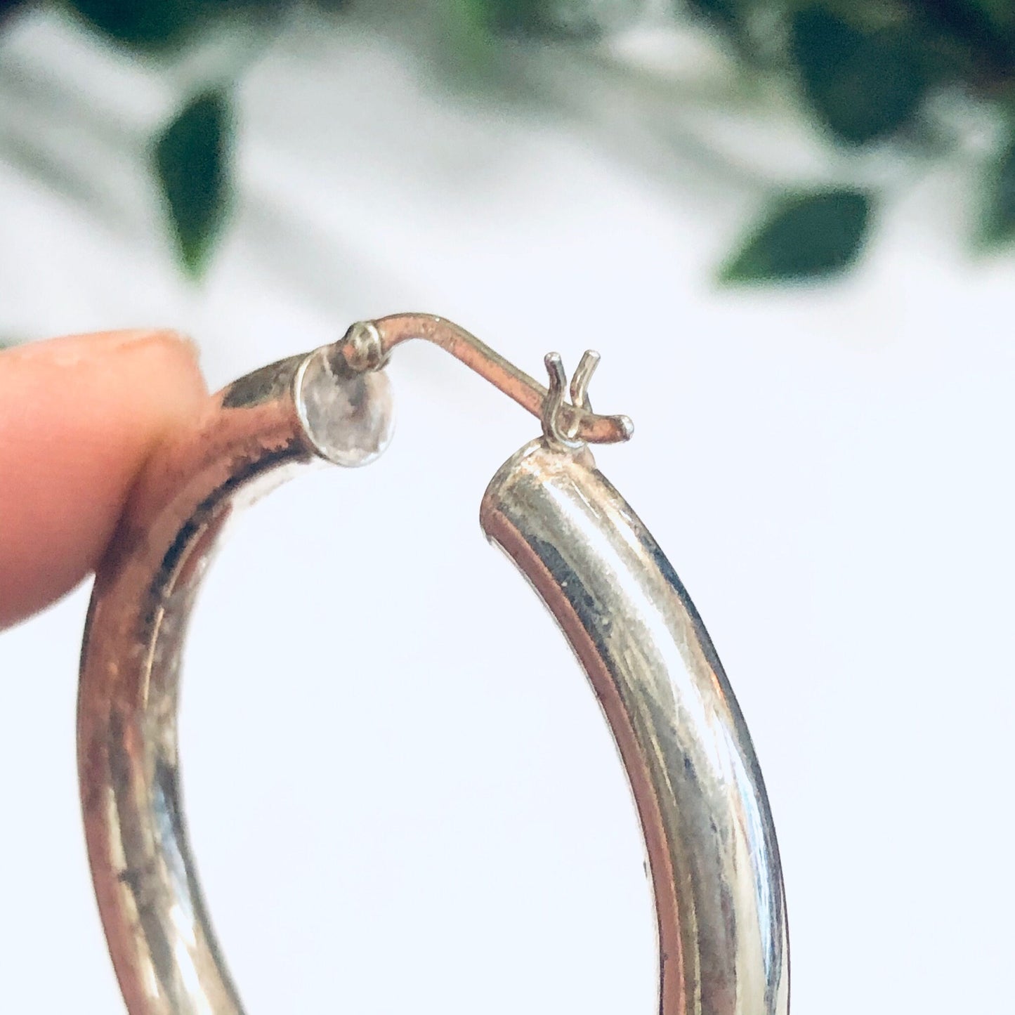 Silver hoop earrings held by a hand with green leaves in the background, medium sized hollow silver hoops for pierced ears