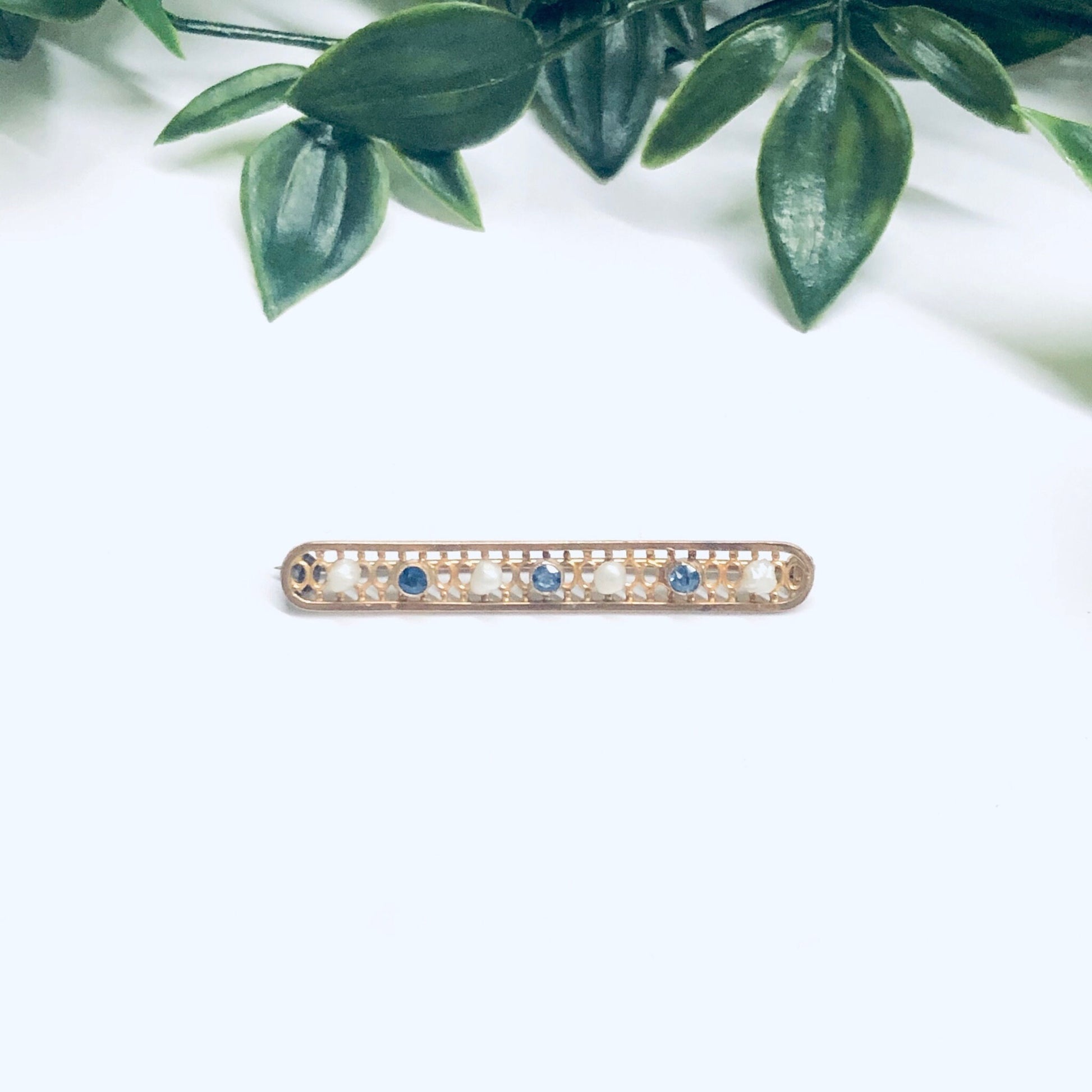 Vintage 14K yellow gold lattice bar pin with seed pearls and faceted blue gemstones, measuring 1 3/4 inches long, on a white background with green leaves above.