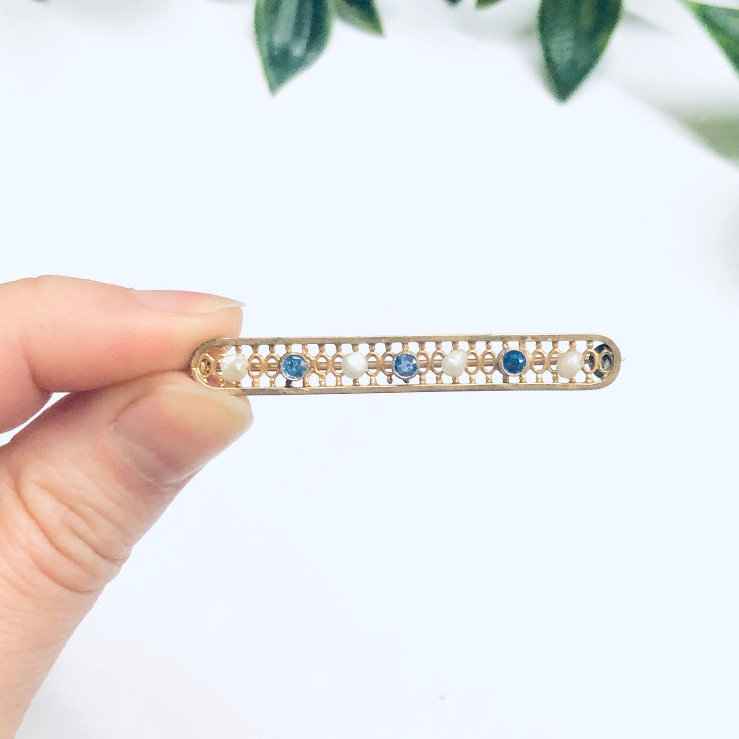 Vintage 14K yellow gold bar pin brooch featuring intricate lattice design with pearls and faceted blue gemstones, measuring 1 3/4 inches long, held in hand with greenery in background.