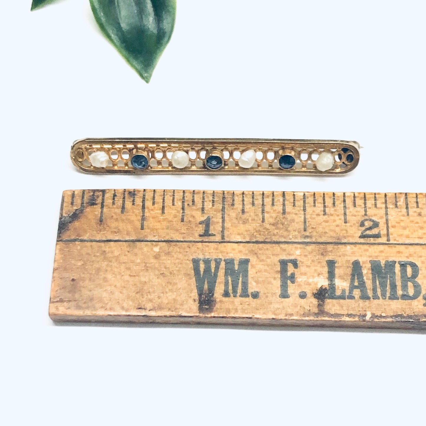 Vintage 14K yellow gold bar pin with pearl seed and faceted blue gemstones in a lattice design, measuring 1 3/4 inches long, shown next to a wooden ruler with the name "Wm. F. Lamb" printed on it, against a white background with a green leaf in the upper left corner.