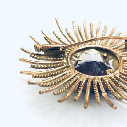 Vintage sunburst brooch with rose gold rays and a round alexandrite gemstone that appears purple and blue, isolated on a white background