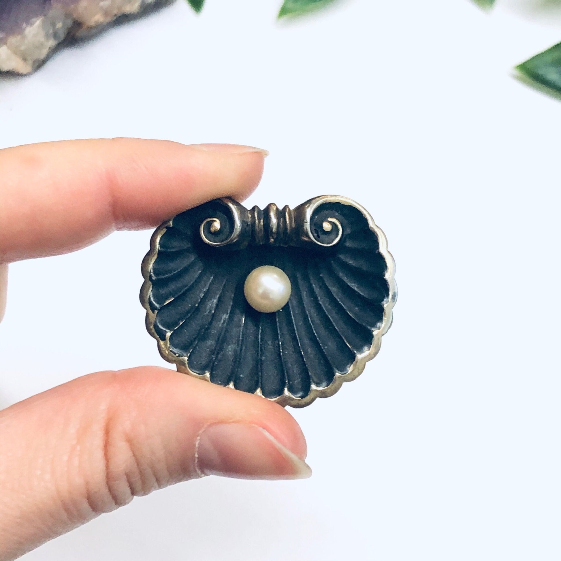 Black seashell-shaped brooch with gold accents and a white pearl center, held between a person's fingers against a white background with leaves.