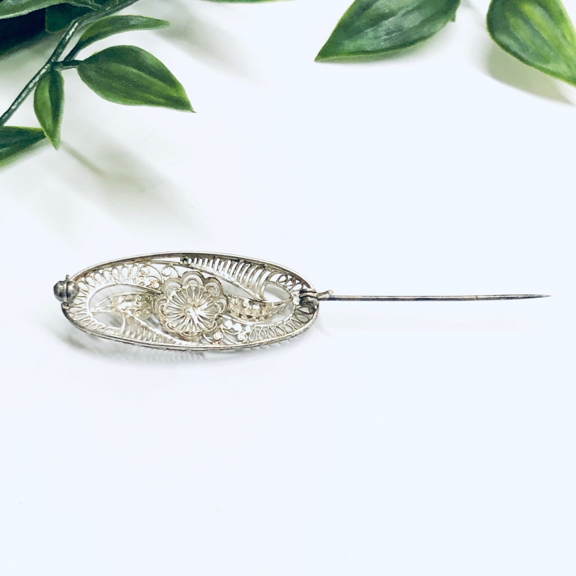 Vintage silver brooch with intricate flower design and cut-out details, delicate silver pin, ornate vintage jewelry piece, displayed on white surface with green leaves in background