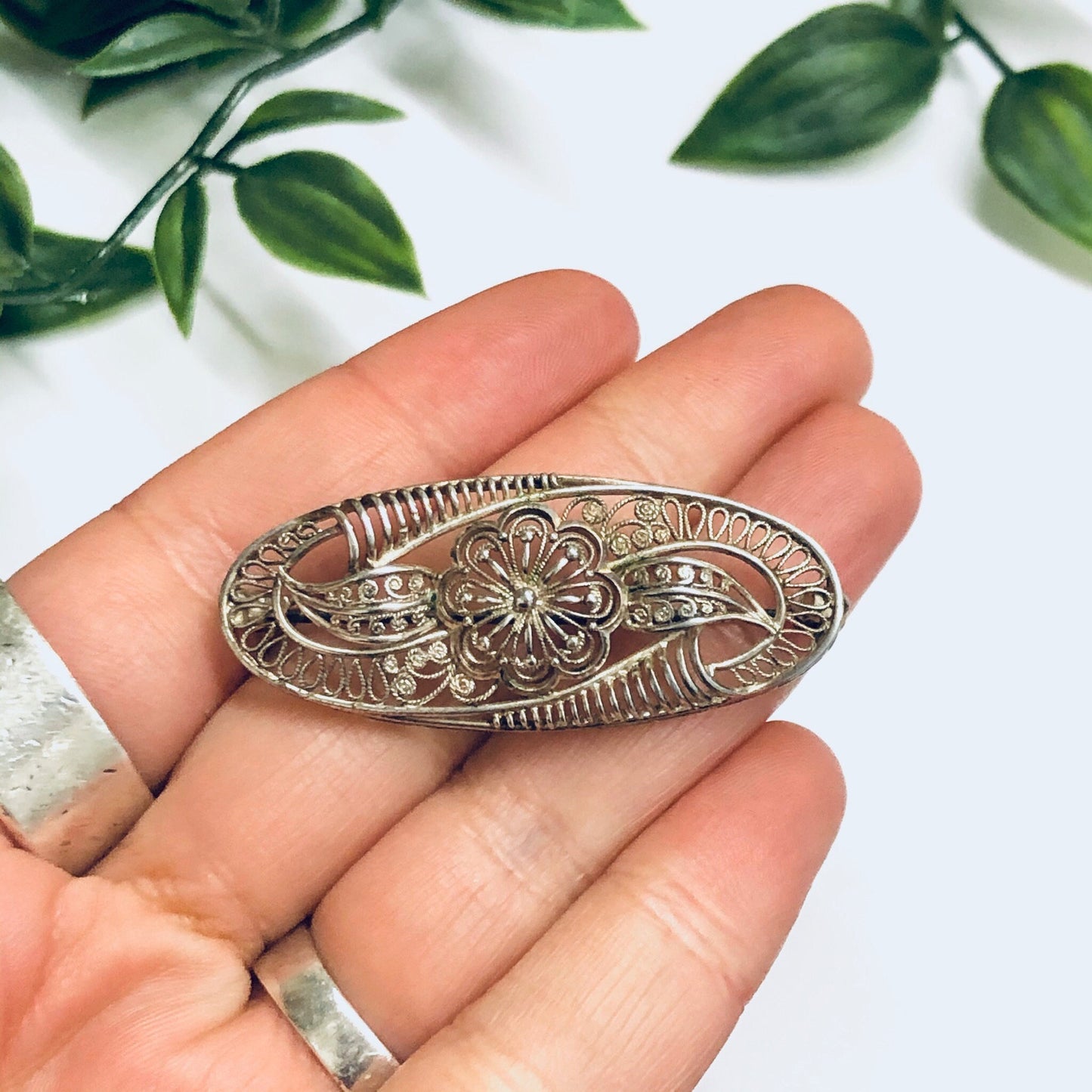 Vintage silver brooch with intricate flower design featuring cutout details, held in palm of hand with green leaves in soft focus background