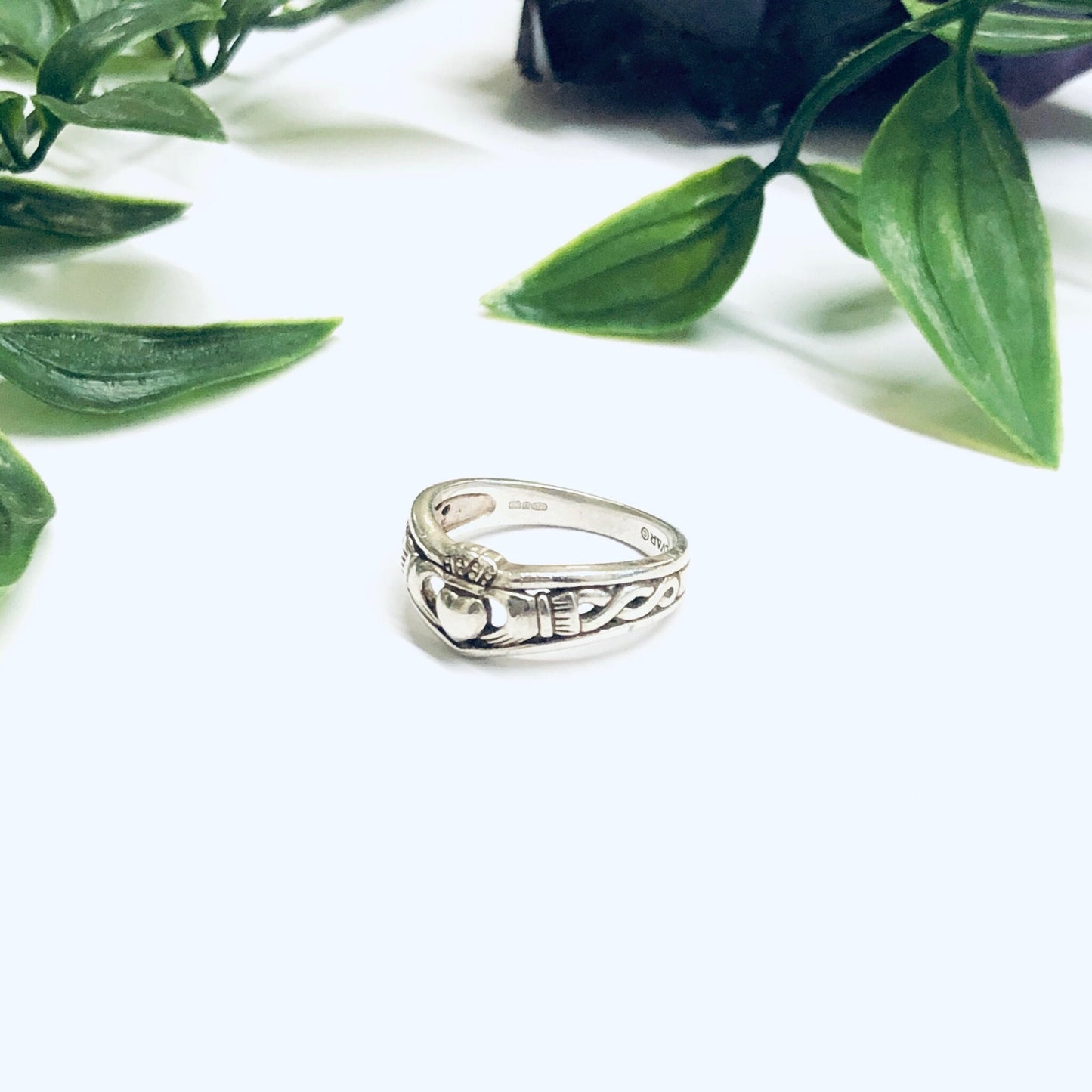 Sterling silver Claddagh ring with heart-shaped design surrounded by lush green leaves on a white background, symbolizing love, loyalty and friendship in Irish tradition.