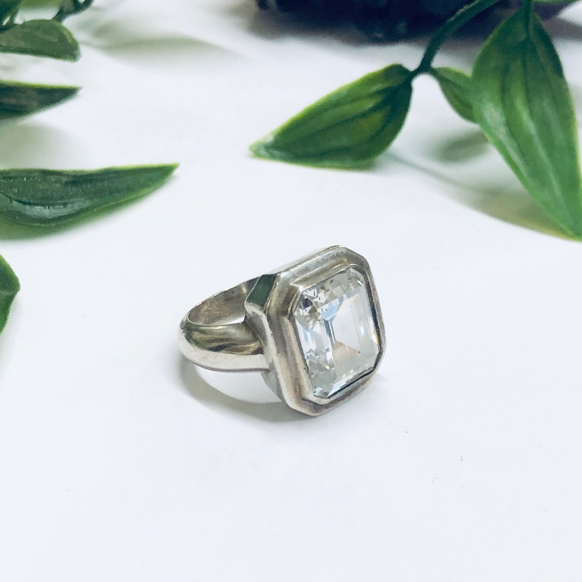 Large silver cocktail ring with clear rectangular gemstone set against green leaves, unique vintage statement jewelry that could serve as a promise ring.