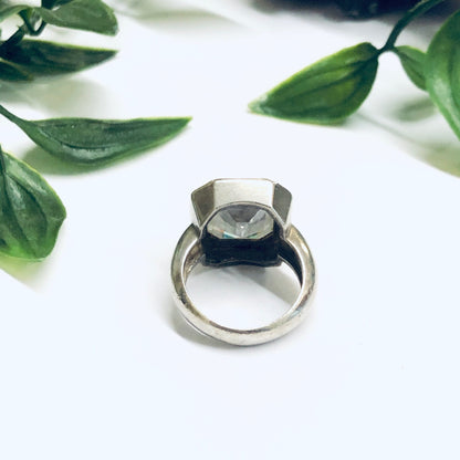 Large silver statement cocktail ring with unique geometric design and dark blue stone, surrounded by green leaves on a white background.