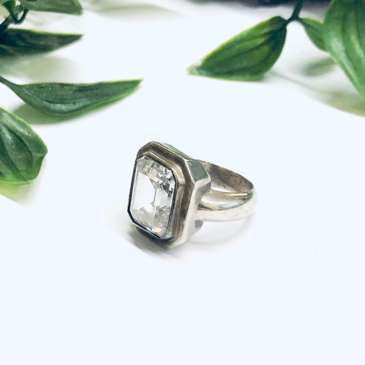 Silver statement cocktail ring with rectangular crystal set among green leaves on white background