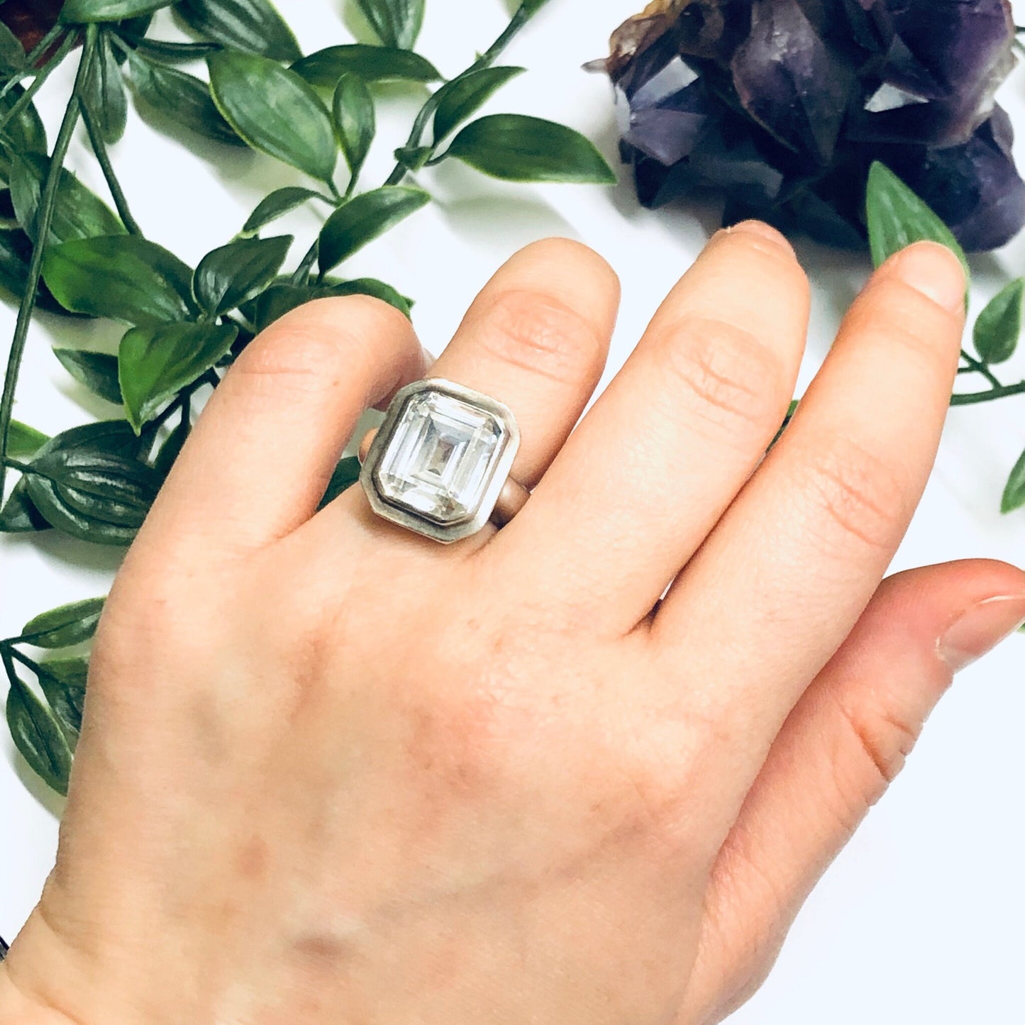 Large silver cocktail ring with unique geometric design worn on hand, displayed with lush green foliage in background.