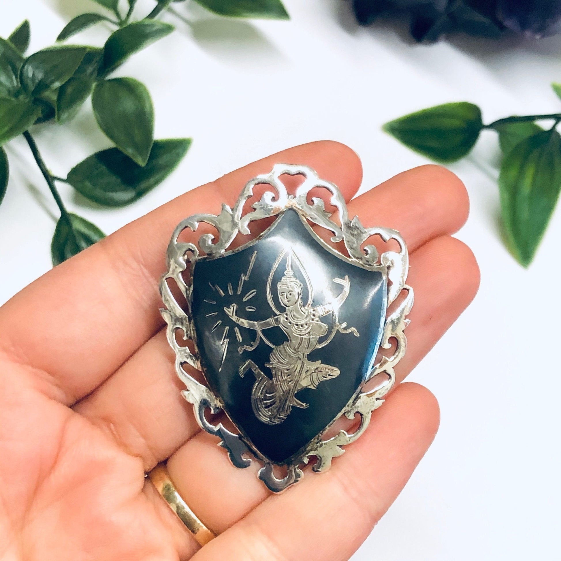 Vintage Siam sterling silver brooch depicting the Goddess of Lightning Mekkala, crafted by Amfarco. The intricate silver pin features an engraved design on a dark blue enamel background, held in a person's hand with greenery in the background.