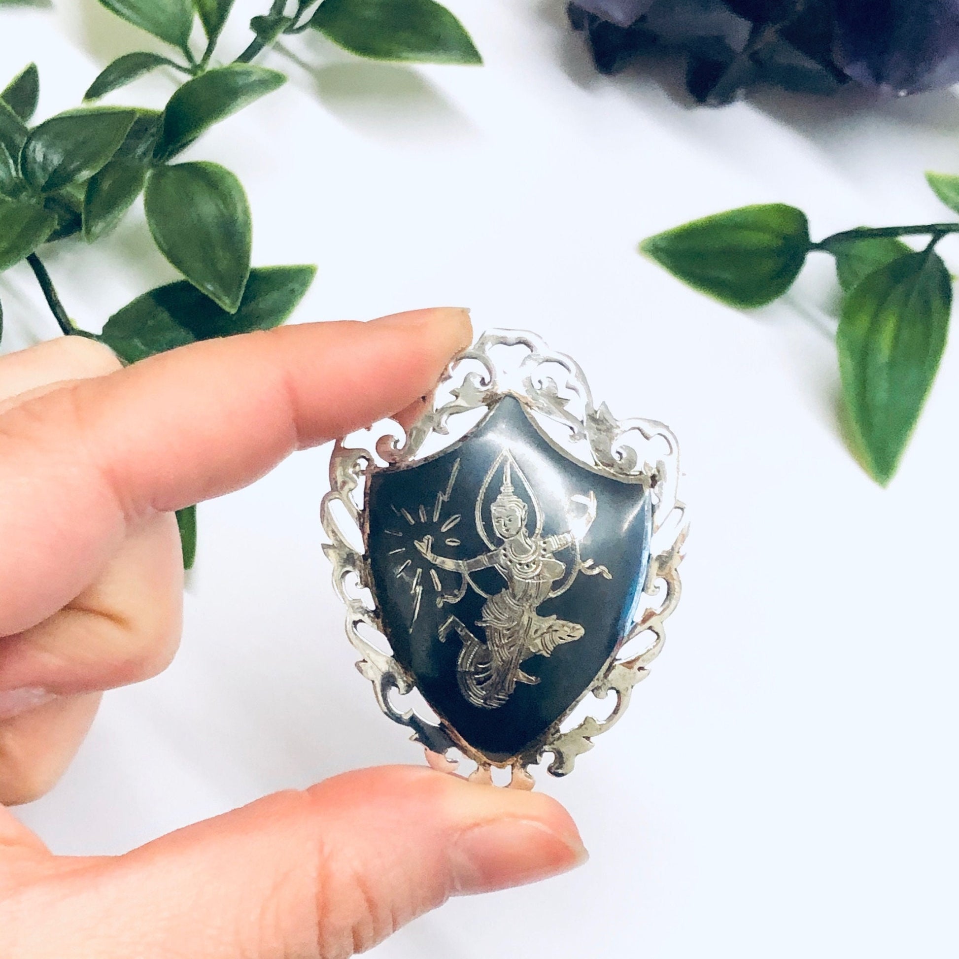 Vintage Siam sterling silver brooch depicting the Goddess of Lightning Mekkala, crafted by Amfarco. The intricate brooch features an engraved floral design against a dark blue enamel background, held delicately in a hand against a white surface with green foliage.