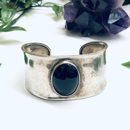 Vintage silver cuff bracelet featuring a large oval onyx stone in the center, set against a white background with purple flowers.