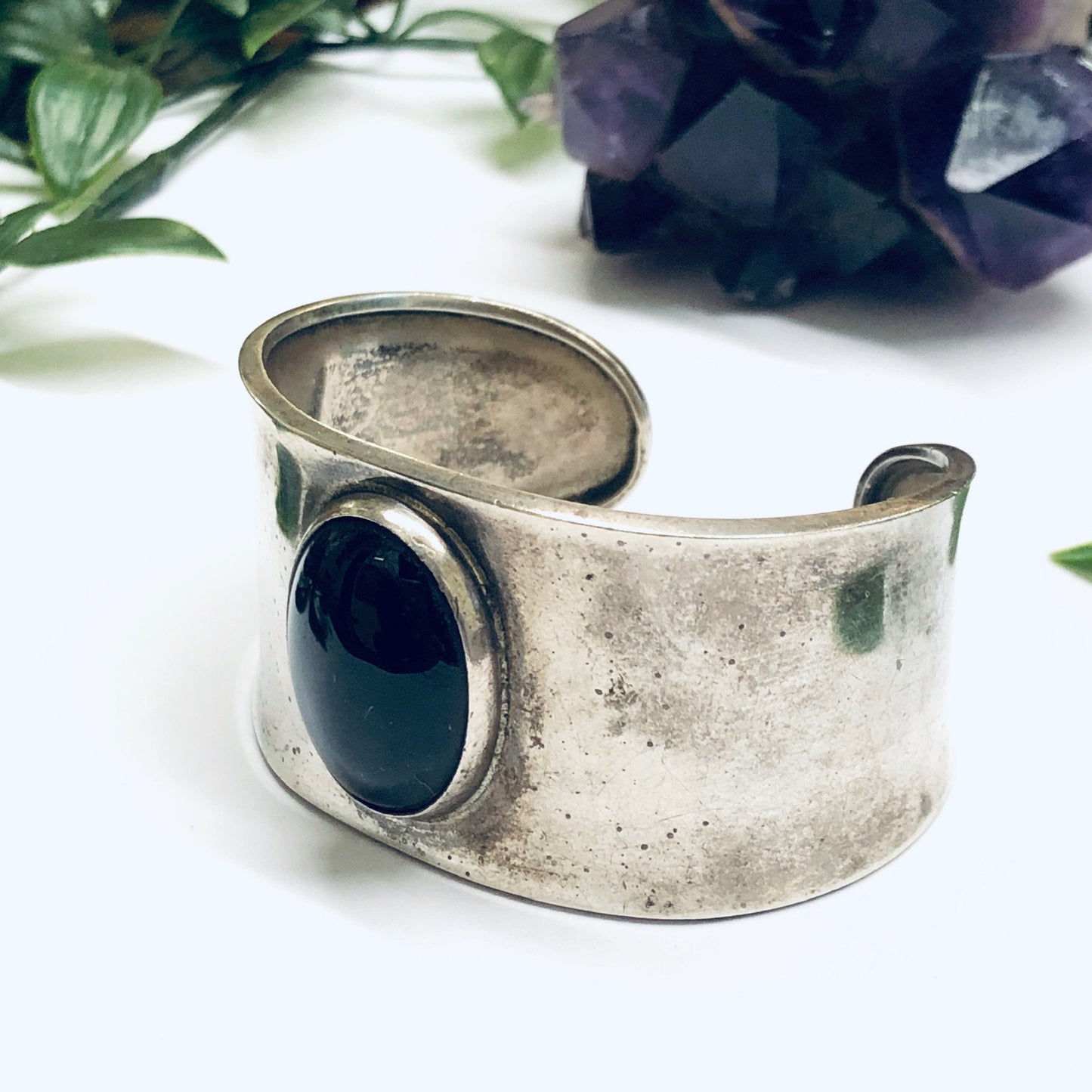 Vintage silver cuff bracelet with oval onyx cabochon stone set in bezel, worn antique finish, shown with dark purple amethyst crystal in soft white background.