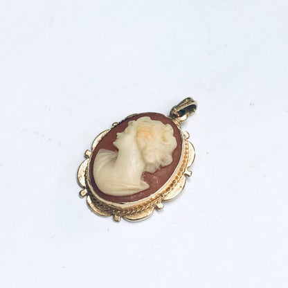 Vintage 14K gold pendant featuring a small, intricately carved cameo depicting a woman's profile against a rust-colored background, set in an ornate yellow gold frame with delicate openwork and scroll detailing.