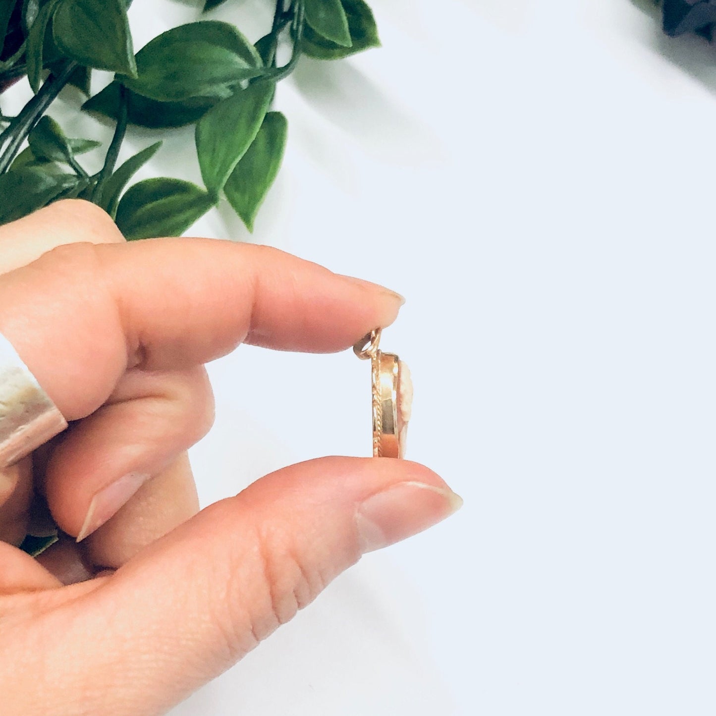 A hand holding a small vintage 14K yellow gold cameo pendant against a white background with a green plant visible.