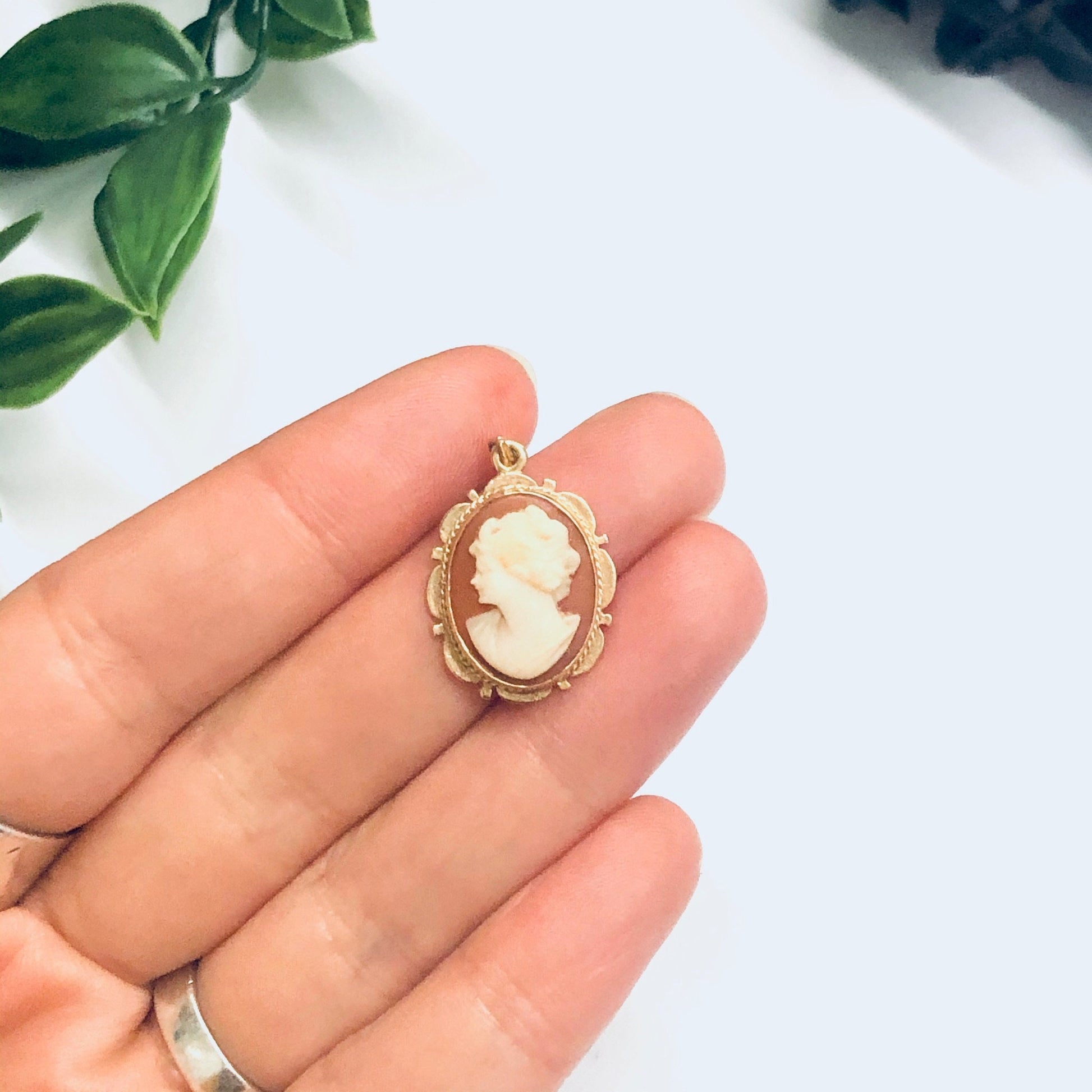 Small vintage 14K yellow gold cameo pendant held in a hand against a white background with green foliage.