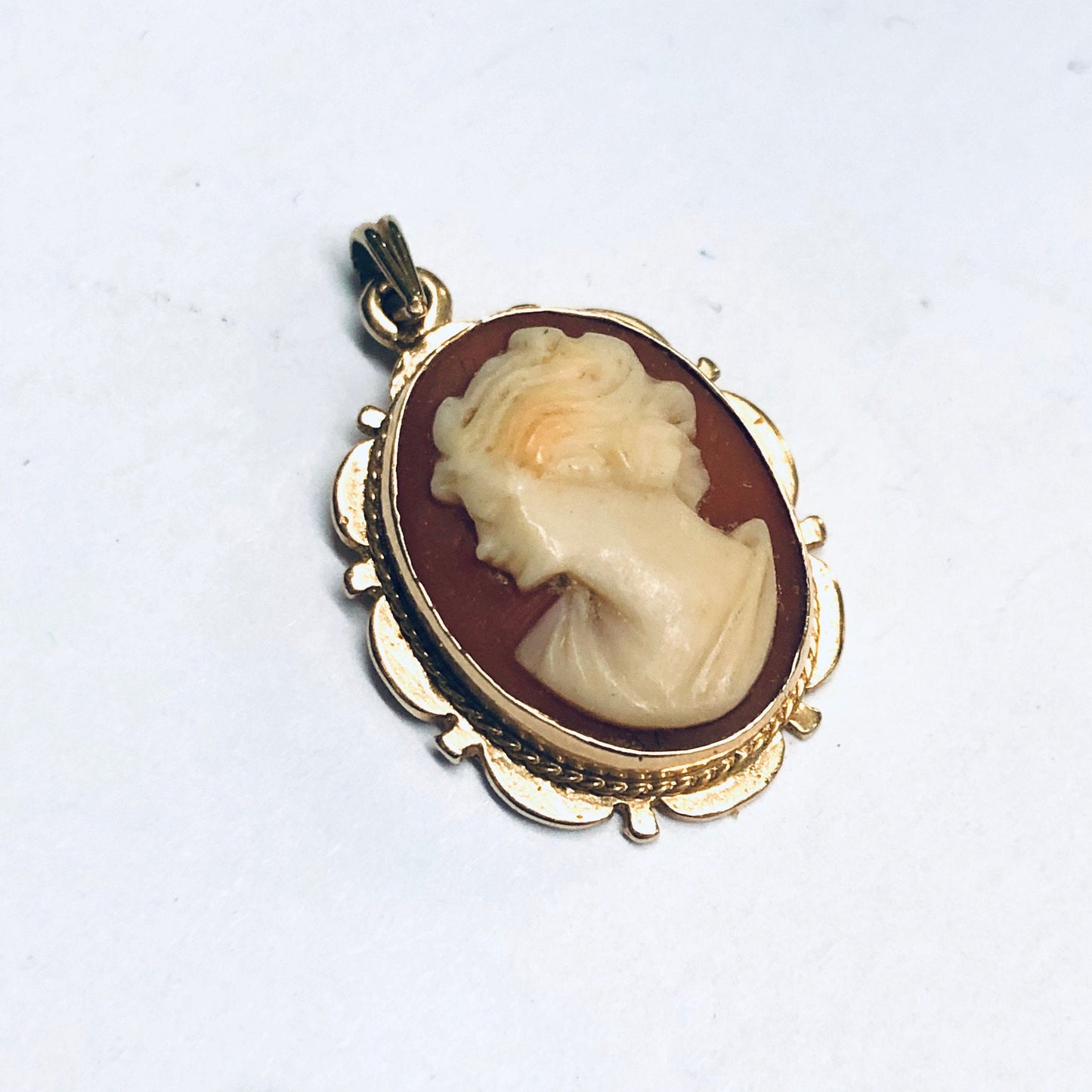 Vintage 14K yellow gold oval pendant featuring a carved shell cameo depicting a woman's profile set in an ornate scalloped frame against a dark red background.