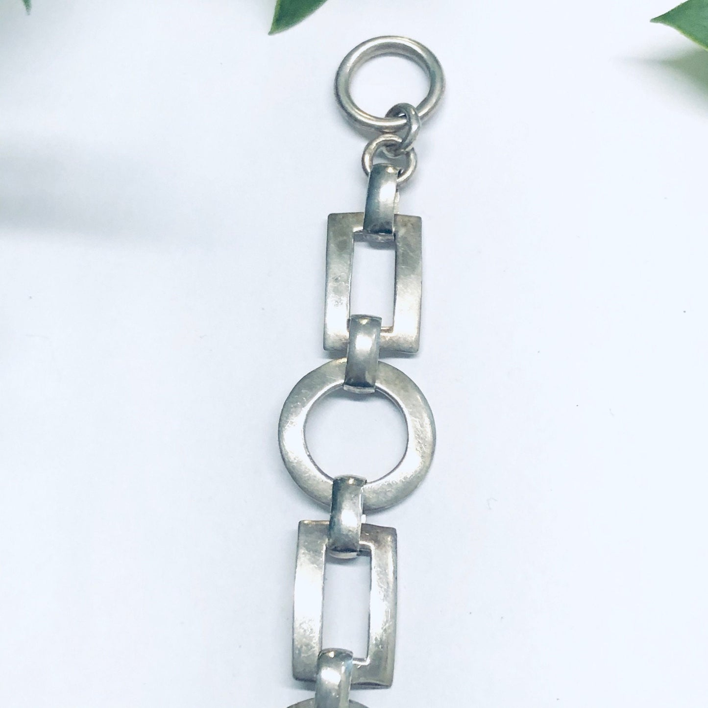 Silver geometric link bracelet with circular and rectangular cutout designs, connected by a toggle closure clasp, shown against a white background with leaves.