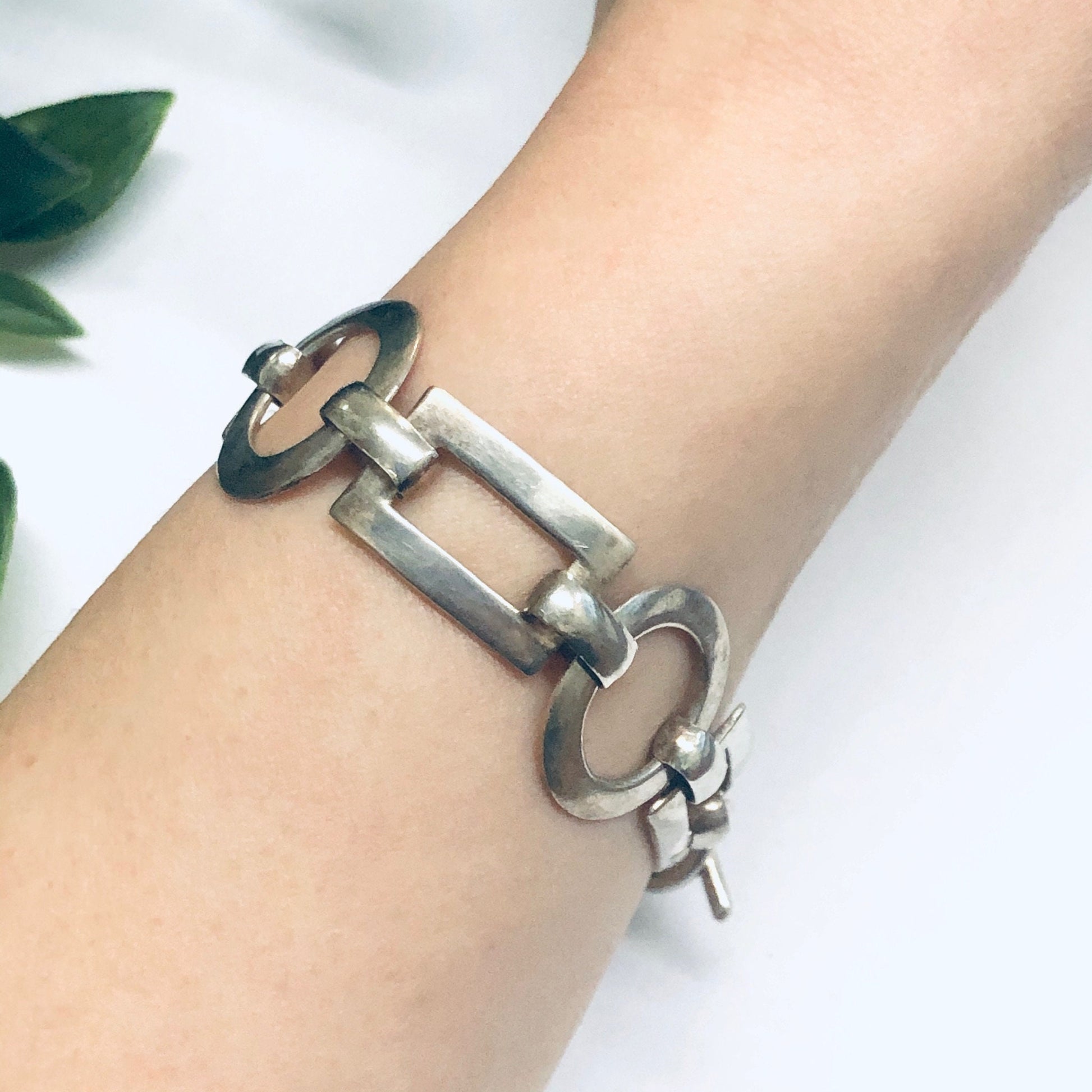 Geometric silver link bracelet with toggle closure, featuring cutout circles and rectangles, on wrist with plant in background