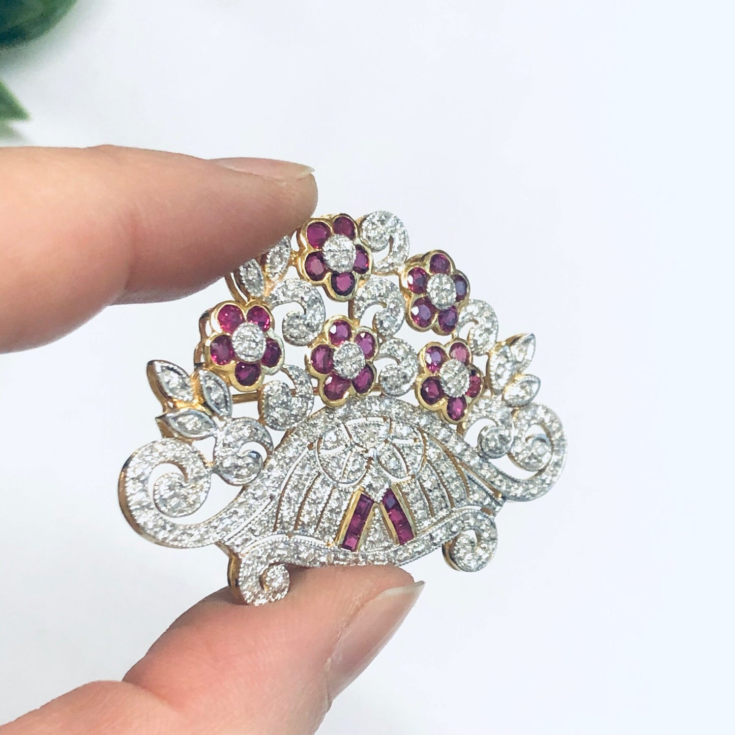 Vintage 18K yellow gold brooch pin featuring a floral design adorned with rubies and diamonds, held in a person's fingers against a white background.