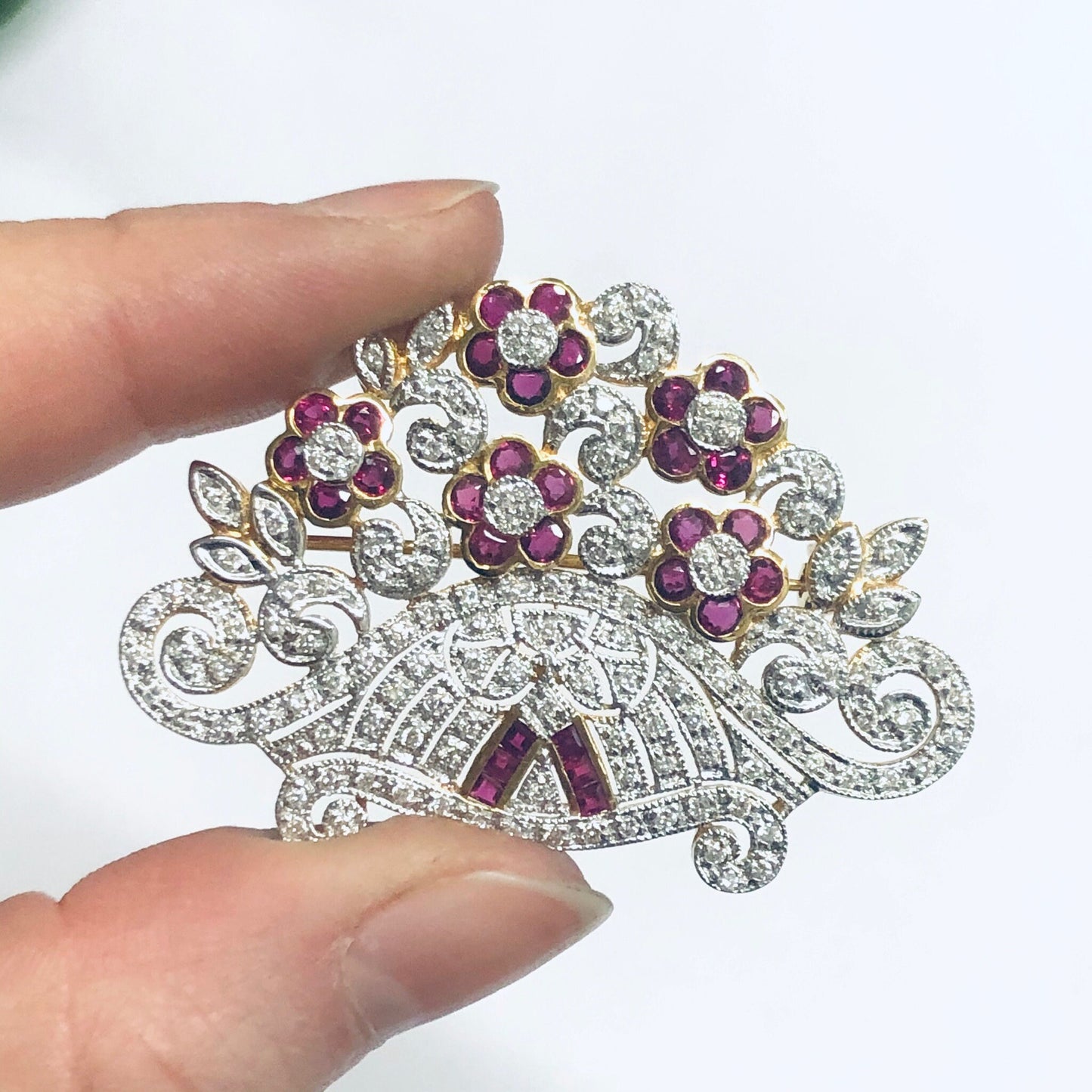 Vintage 18K yellow gold brooch pin with rubies and diamonds in an intricate floral design, held in fingers against a white background.