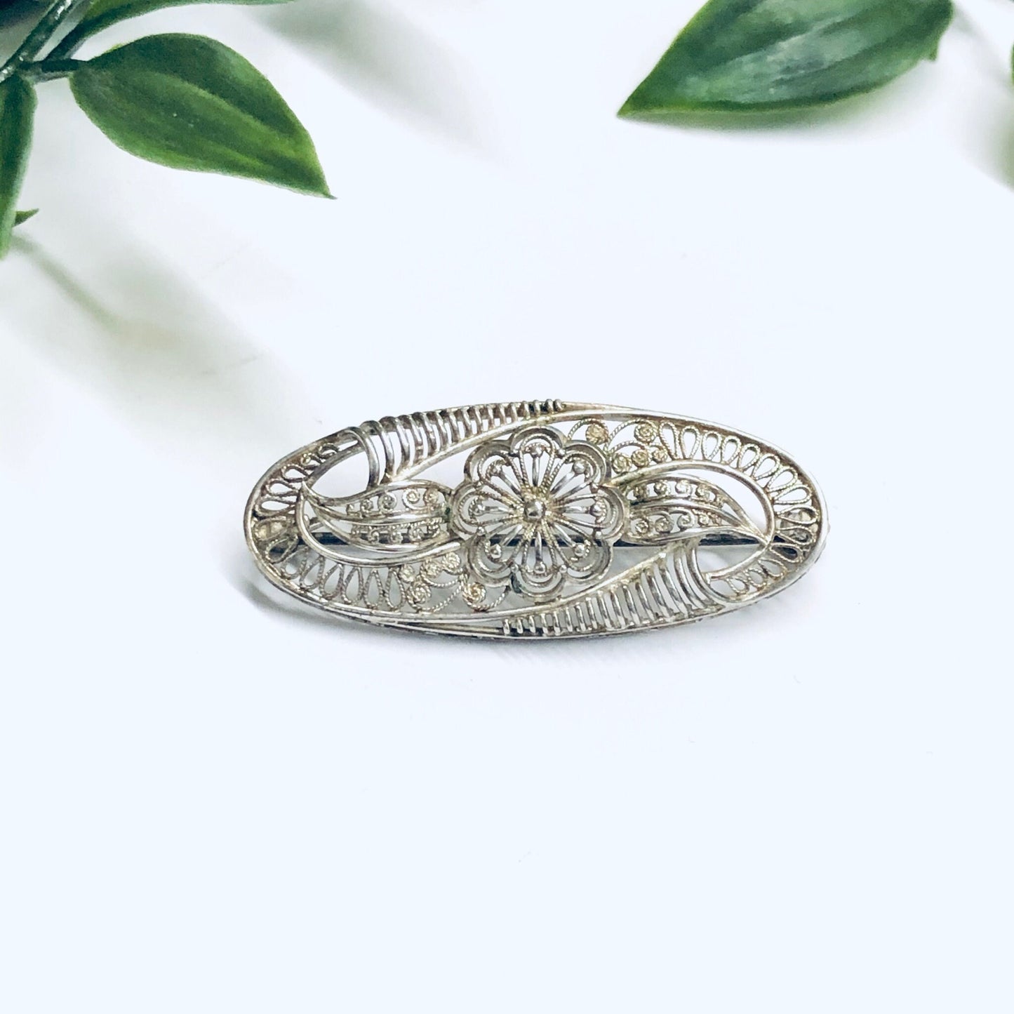 Vintage silver brooch featuring an ornate cut-out flower design, displayed on a white surface with green leaves in the background, creating a delicate and elegant piece of antique jewelry.