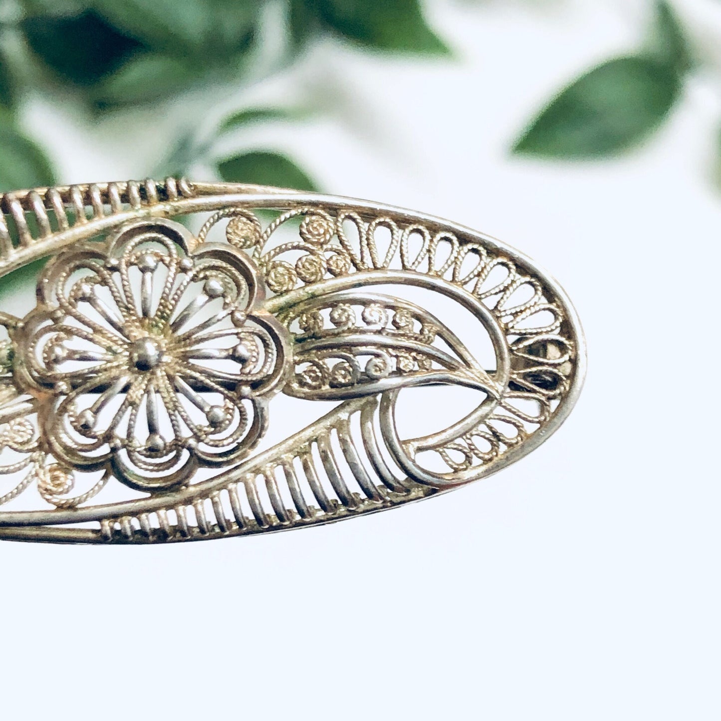 Vintage silver brooch with intricate flower and leaf cut-out design, delicate pin clasp visible on back, set against soft blurred green foliage background.