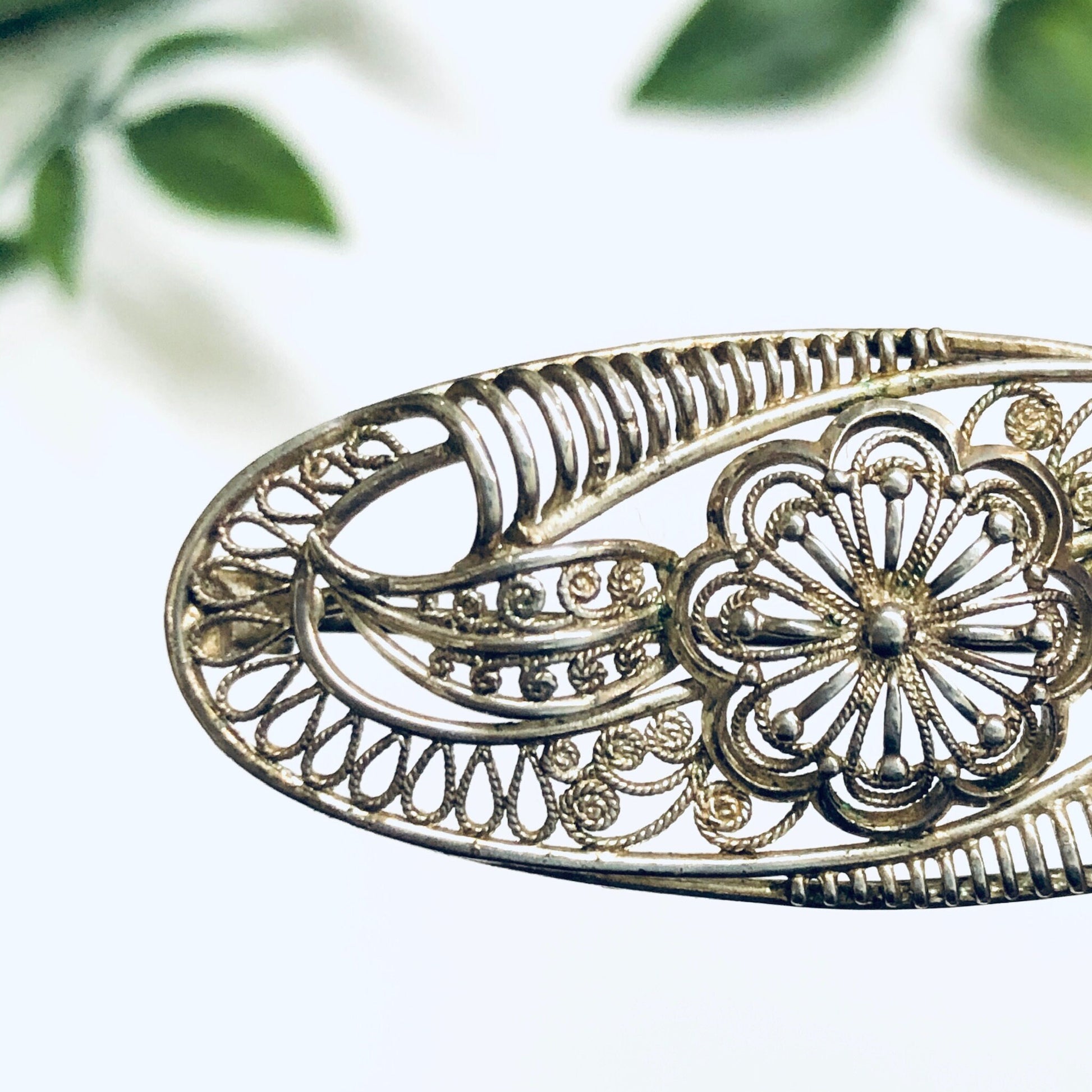 Vintage silver brooch with ornate flower and cutout design, delicate antique pin showcasing intricate metalwork and floral motif against blurred greenery background.