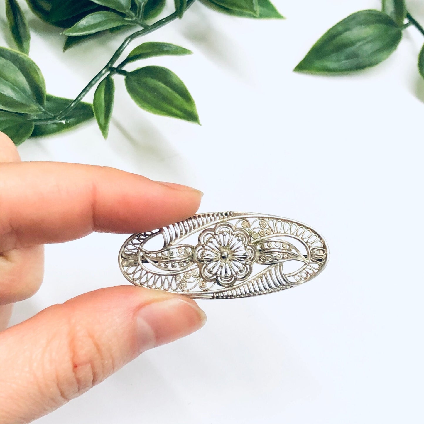 Vintage silver brooch with intricate floral cut-out design, held against a white background with green leaves, delicate ornate jewelry pin