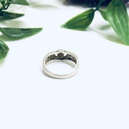 Silver Claddagh ring with three small heart-shaped stones, resting on a white surface with green leaves in the background.