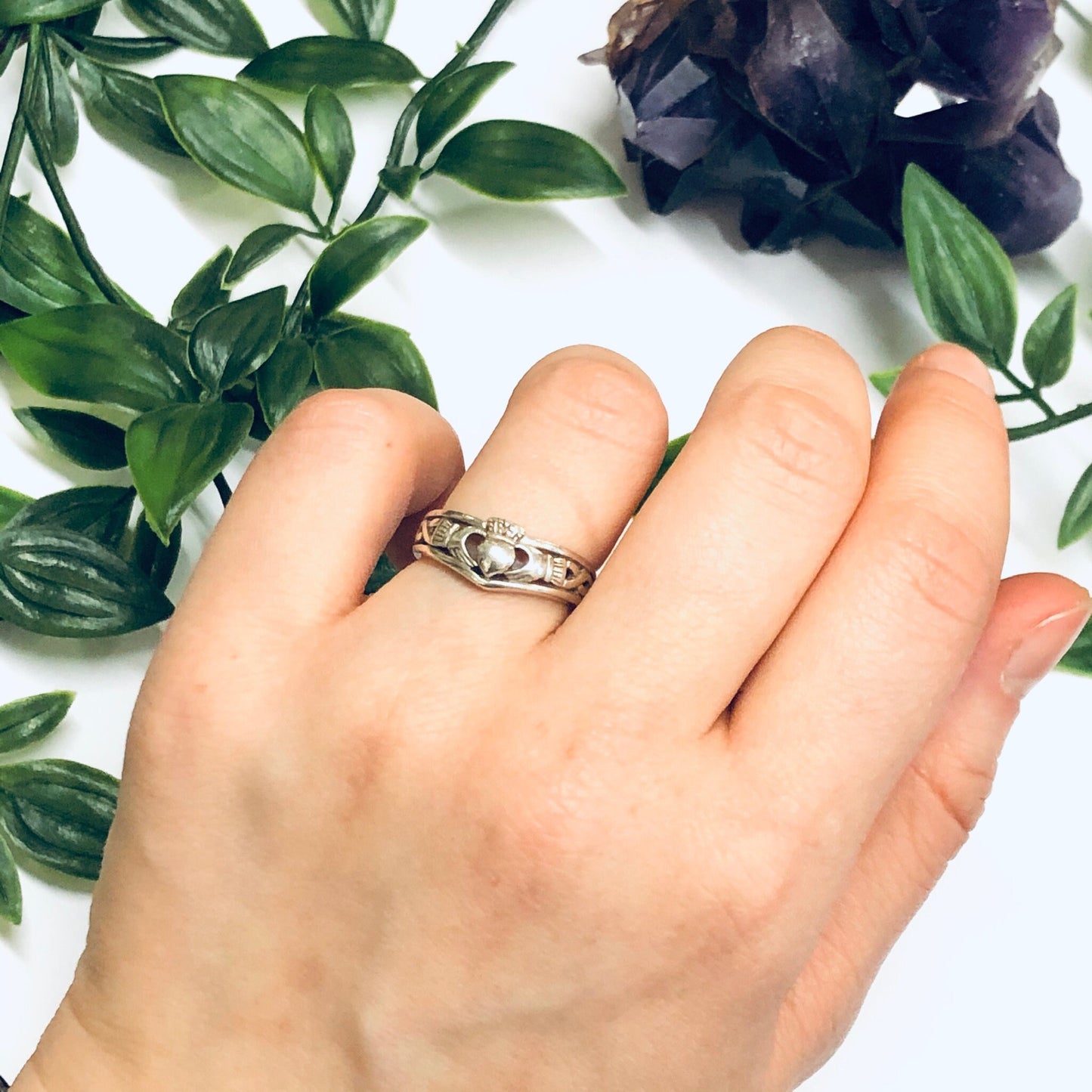 Silver Claddagh ring with heart held by hands, surrounded by green leaves