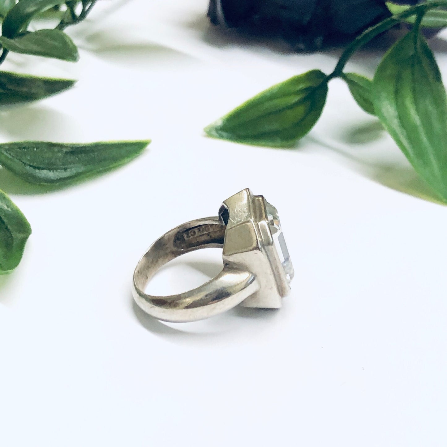 Large vintage silver cocktail statement ring with unique geometric design surrounded by green leaves on white background, promise ring, unique jewelry.