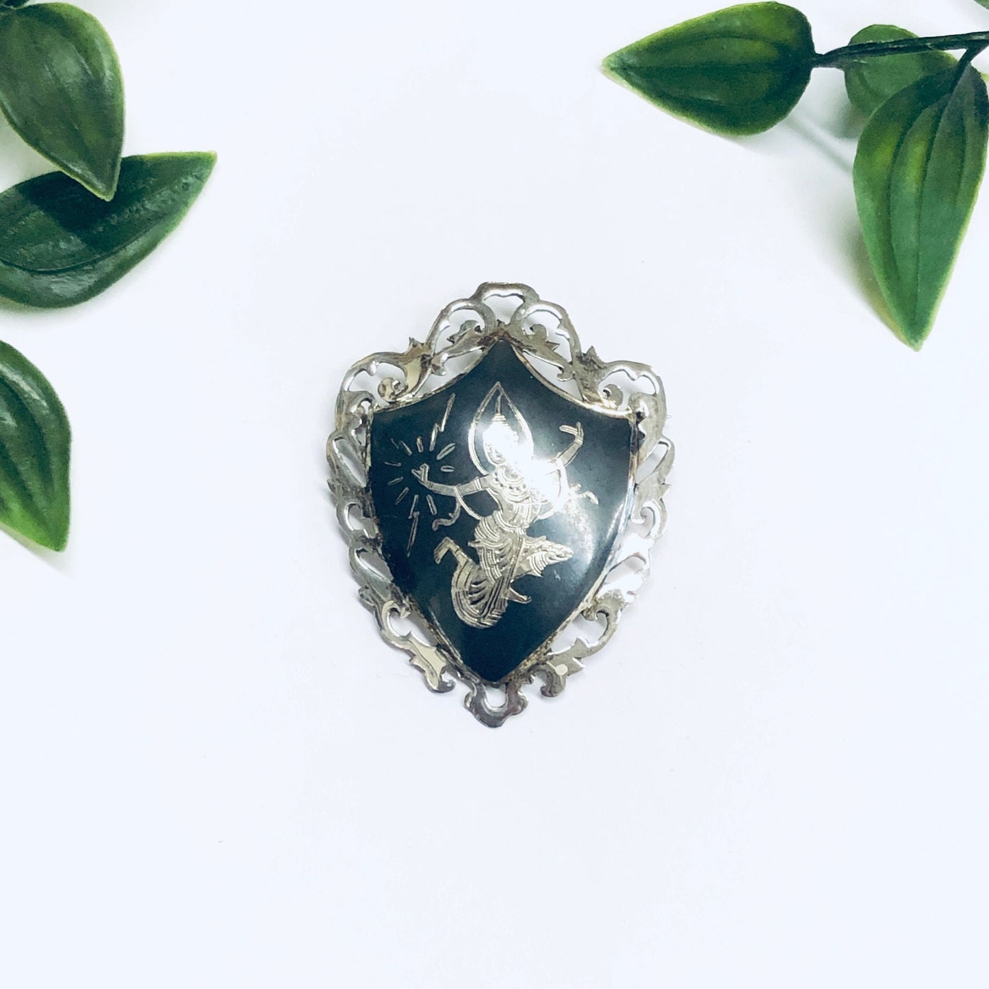 Vintage Siam sterling silver brooch featuring a detailed carving of the Goddess Mekkala, adorned with intricate floral designs around the edges, photographed on a white background with green leaves.