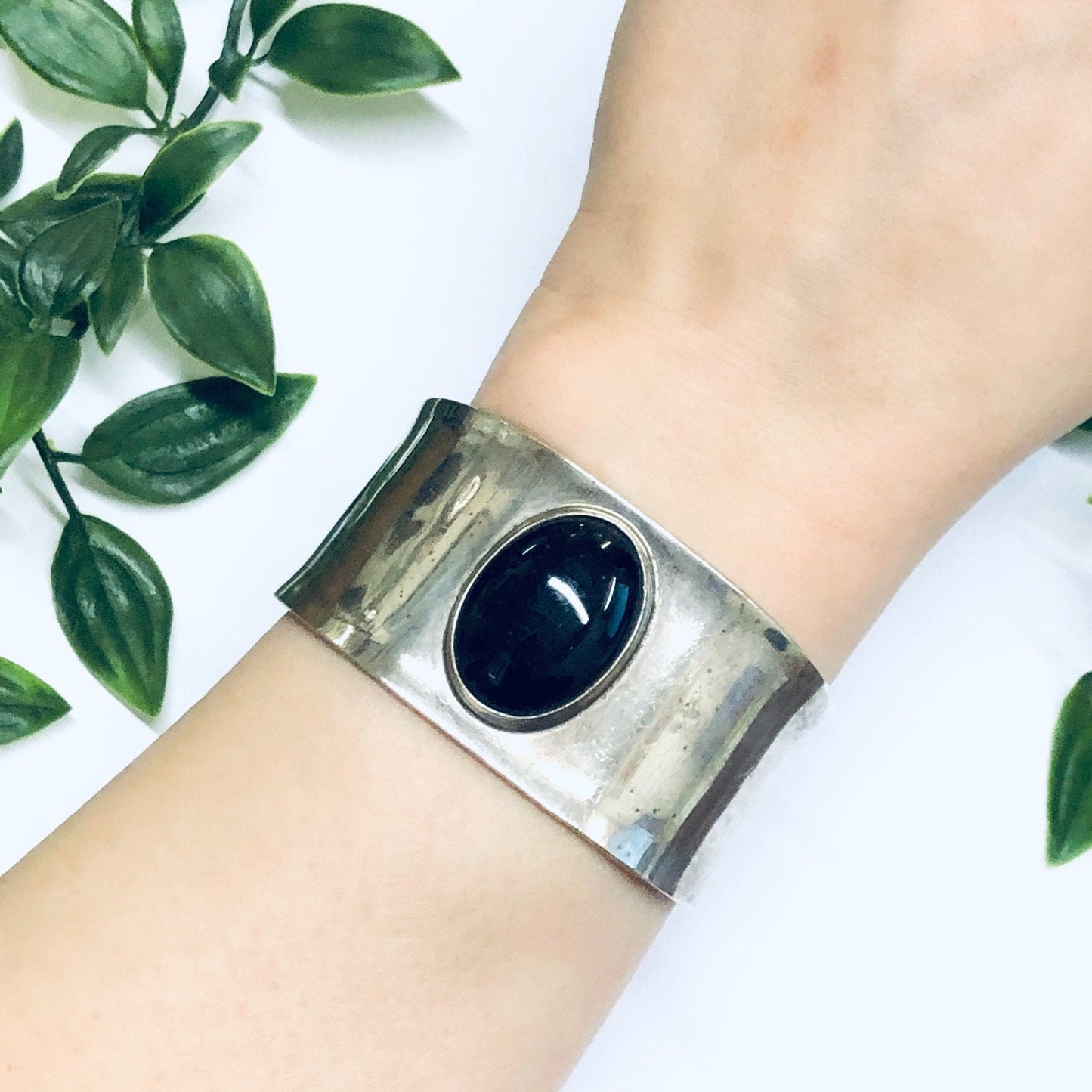 Vintage silver cuff bracelet with large oval onyx stone on female wrist, surrounded by green leaves against white background