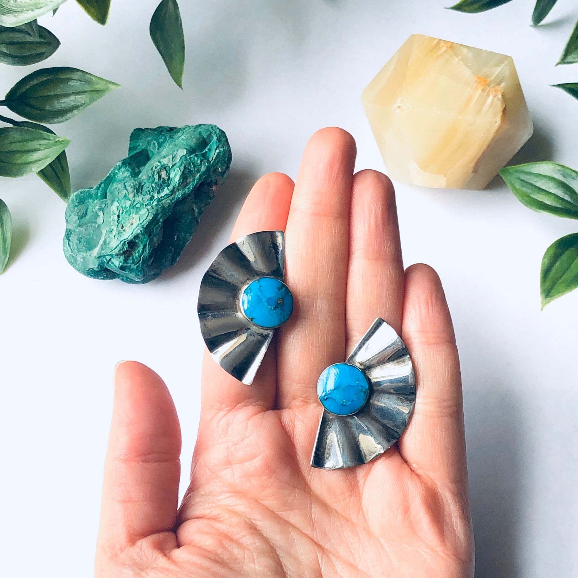 Hand holding vintage silver fan-shaped earrings with blue gemstones, surrounded by green leaves on a white background.