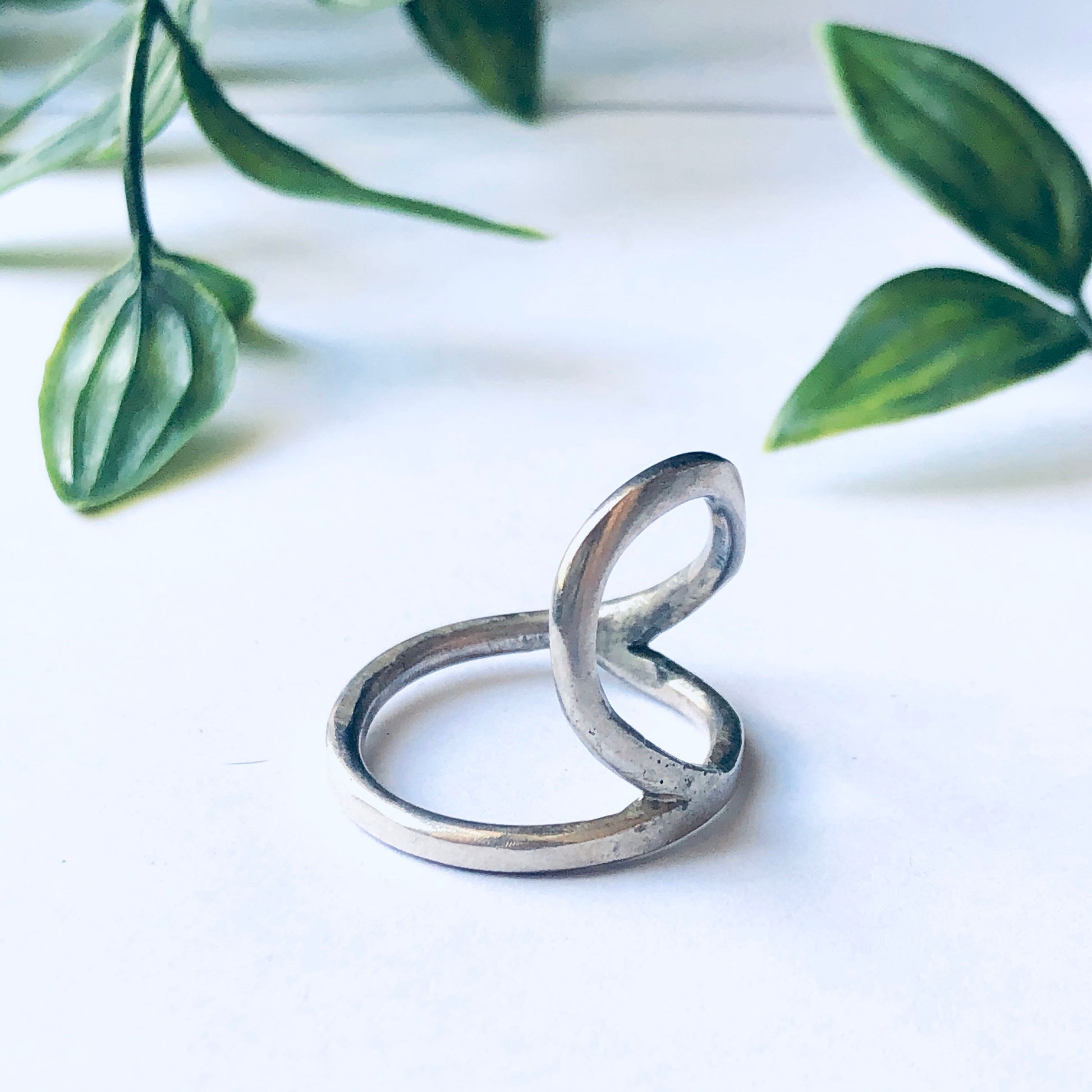 Vintage silver cut out ring with large minimalist abstract design displayed against a white background with green leaves.