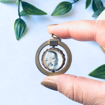 Vintage gold-toned Martin Luther King Jr. spinner pendant with 'I Have a Dream' inscription held between fingers with green leaves in background.
