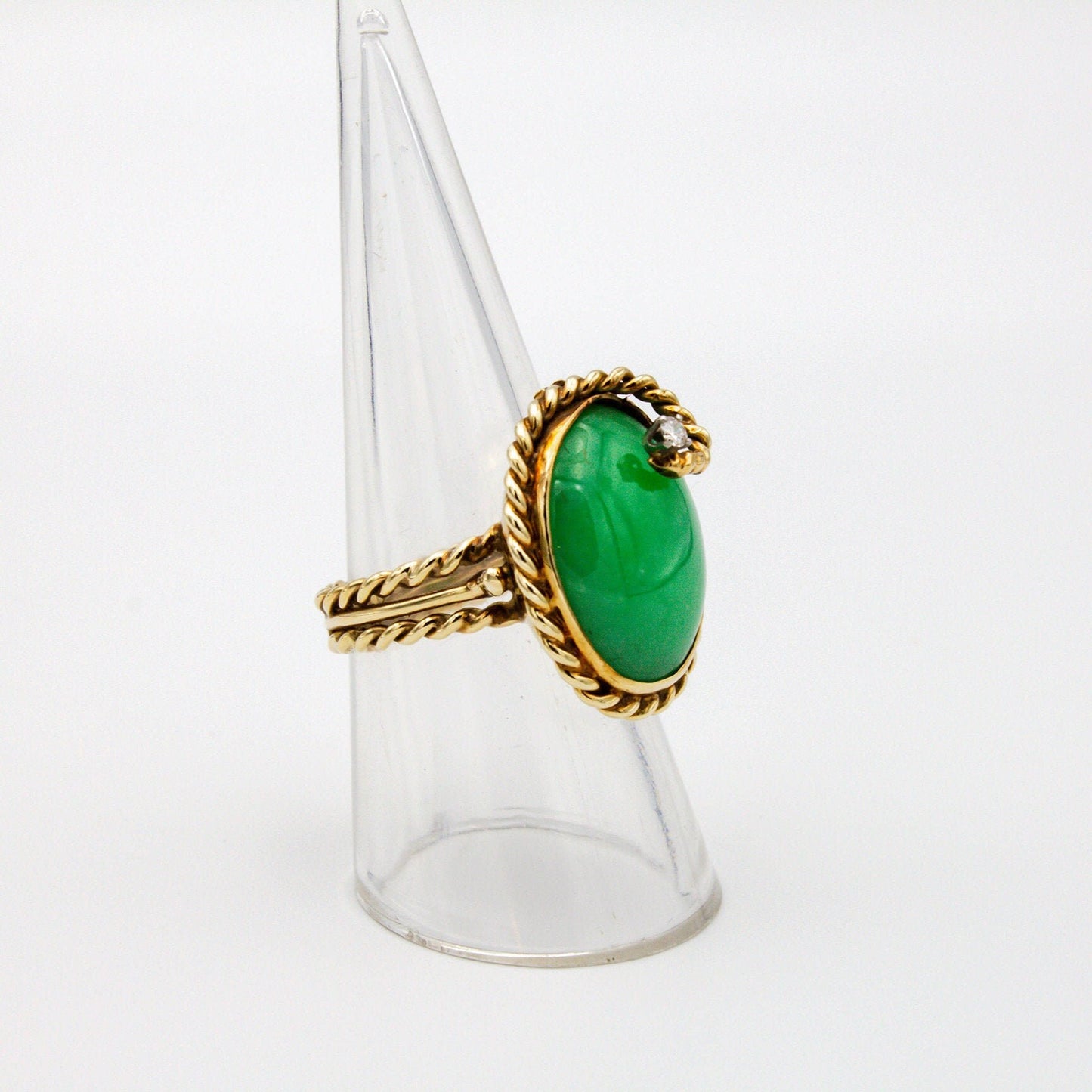 Vintage 14K gold jade and diamond cocktail ring featuring a large oval green jade stone accented by a small diamond, set in a twisting gold band, displayed on a clear ring holder stand.