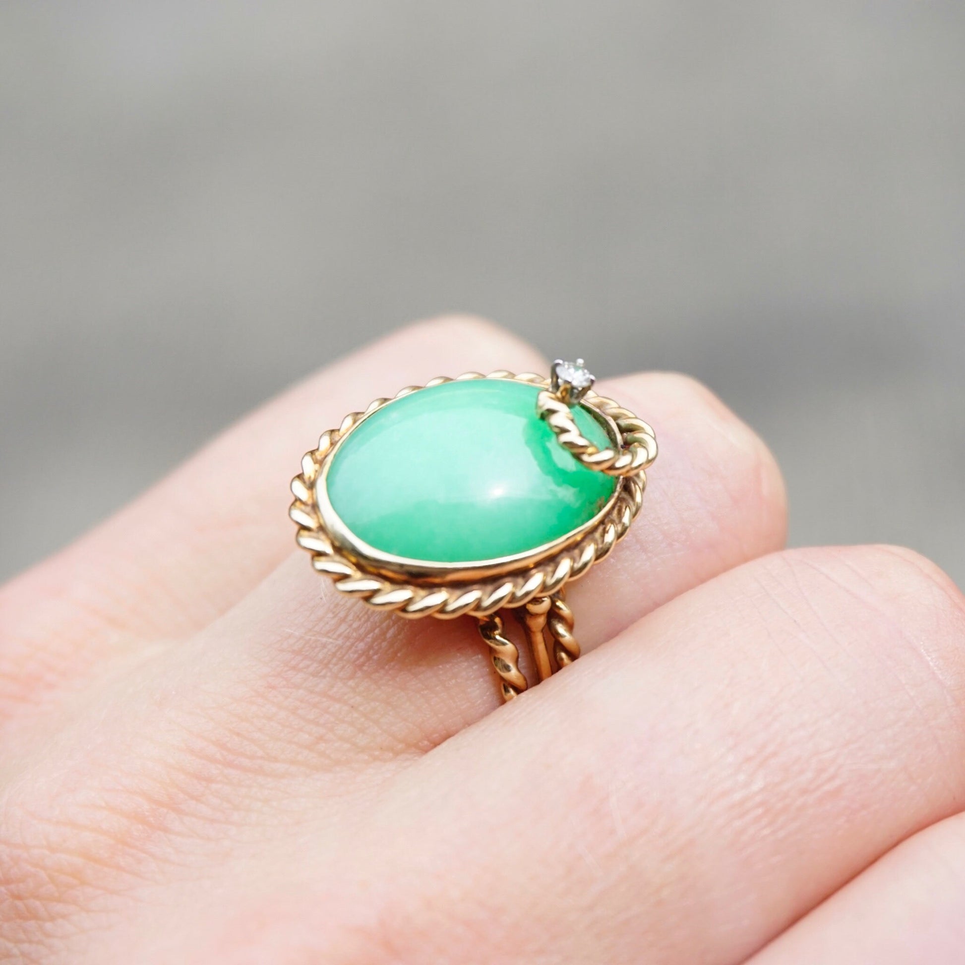 Vintage 14K gold cocktail ring featuring a large oval green jade stone surrounded by a twisted gold band, accented with a single small diamond. The ring is shown worn on a finger against a plain gray background.