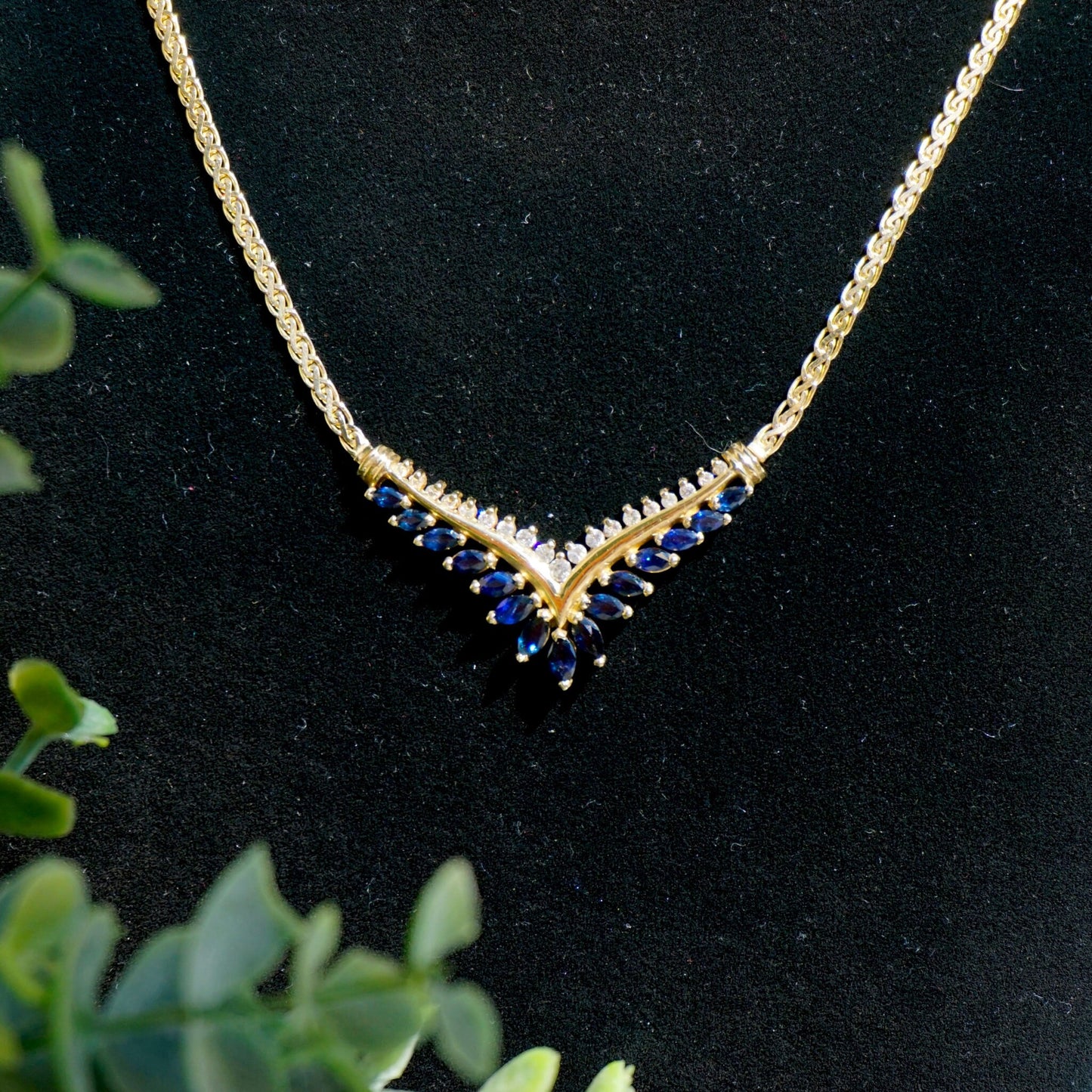 Vintage 14k gold choker necklace featuring elegant sapphire and diamond accents, set in a V-shaped design against a black background with green foliage.